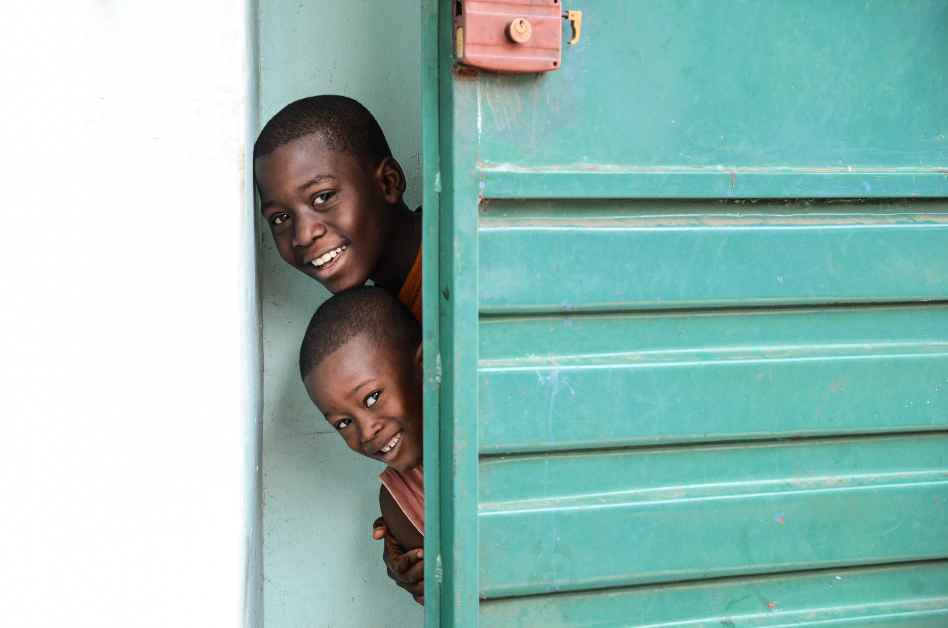 Zwei Kinder, Patenkinder der SOS-Kinderdörfer weltweit, blicken hinter einer grünen Tür hervor und lachen in die Kamera.